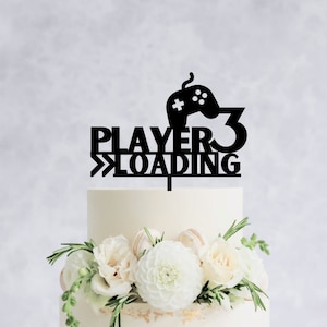 May include: A two-tiered white wedding cake with white flowers and greenery. The cake topper is black and says "Player 3 Loading" with a video game controller icon.