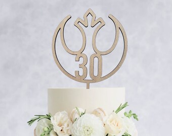 May include: A two-tiered white wedding cake decorated with white flowers and greenery. A wooden cake topper in the shape of the Rebel Alliance symbol with the number 30 in the center.