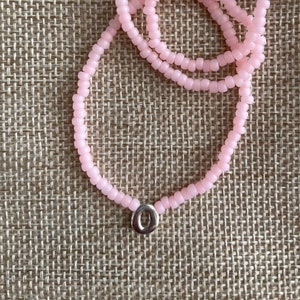 May include: A pink beaded necklace with a silver charm in the shape of the letter "O".