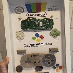 Könnte beinhalten: Gerahmte Präsentation mit Nintendo Super Famicom Controller-Komponenten. Die Anzeige umfasst eine Leiterplatte, Knöpfe und einen Controller mit dem Nintendo-Logo und dem Text Super Famicom. Der Rahmen ist weiß.