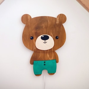 May include: A wooden bear night light with a brown body, a white face, and black eyes. The bear is wearing teal blue pants with two black buttons. The bear is mounted on a white wall.