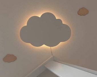 以下が含まれることがあります： 白い壁を暖色系の光で照らす、白い雲の形をしたナイトライト。ライトは壁に取り付けられており、コードが垂れ下がっている。ライトの右側には、壁に小さな木の雲がある。