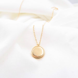Peut inclure: Un collier doré avec un pendentif rond. Le pendentif a un bord perlé et est suspendu à une chaîne délicate. Le collier est présenté sur un tissu blanc texturé.