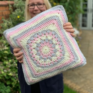 May include: A crocheted cushion with a square design in shades of pink, white, and grey. The cushion is made with a granny square pattern and has a soft, textured look.