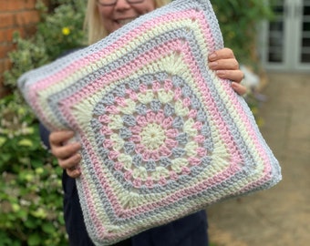 May include: A crocheted pillow with a square design in shades of pink, white, and gray. The pillow is made with a granny square pattern and has a soft, textured look.