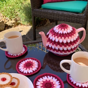 May include: A table setting with a teapot, two mugs, and a plate of mini tarts. The teapot and mugs are on crocheted coasters, and the teapot has a crocheted cover. The table is made of woven wicker.