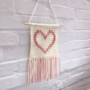 Op de afbeelding: Een witte gehaakte wandhanger met een roze hartontwerp en franjes. Het hart is gemaakt van opgeheven roze garenlussen.