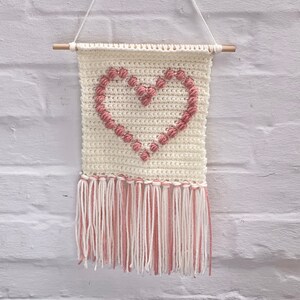Op de afbeelding: Een witte gehaakte wandhanger in de vorm van een hart met roze garen accenten en een franje rand.