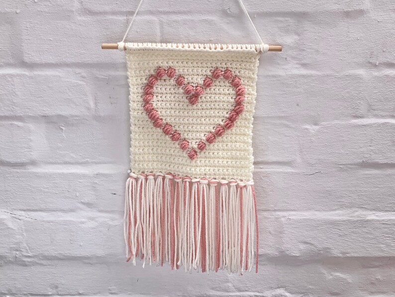 Op de afbeelding: Een witte gehaakte wandhanger in de vorm van een hart met roze garen accenten en een franje rand.