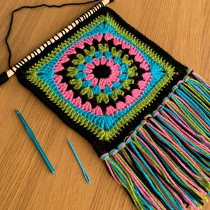 May include: A black crochet wall hanging with a colorful geometric pattern in shades of pink, blue, green, and black. The hanging has a fringe border and is attached to a wooden dowel.