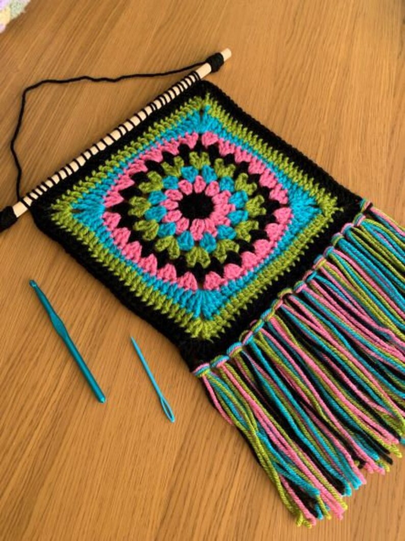 May include: A black crochet wall hanging with a colorful geometric pattern in shades of pink, blue, green, and black. The hanging has a fringe border and is attached to a wooden dowel.