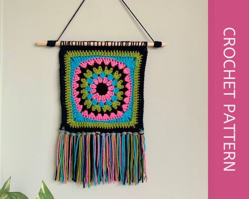 May include: A crocheted wall hanging with a black, green, pink, and blue mandala design. The hanging has a fringe border and is attached to a wooden dowel.