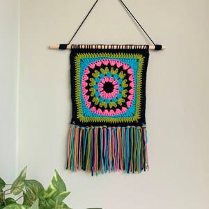 May include: A colorful crocheted wall hanging with a black center and green, pink, and blue accents. The hanging has a fringe border and is attached to a wooden dowel.