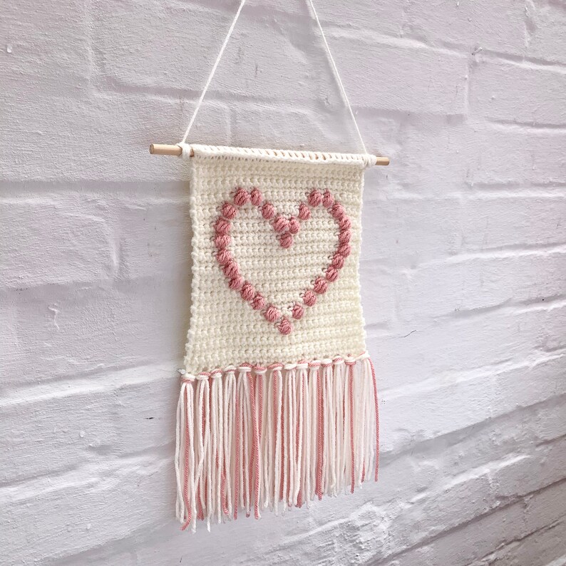 Op de afbeelding: Een witte gehaakte wandhanger met een roze hartontwerp en franjes. Het hart is gemaakt van opgeheven roze garenlussen.