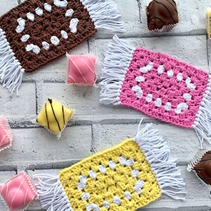 May include: Three crocheted squares with white fringe. Each square has a different colour: brown, pink, and yellow. The squares are arranged on a white brick background with small, colourful pastries.
