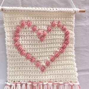 Op de afbeelding: Een witte gehaakte wandhanger met een roze hartontwerp. Het hart is gemaakt van kleine roze garenballen. De hanger heeft witte franjes aan de onderkant.