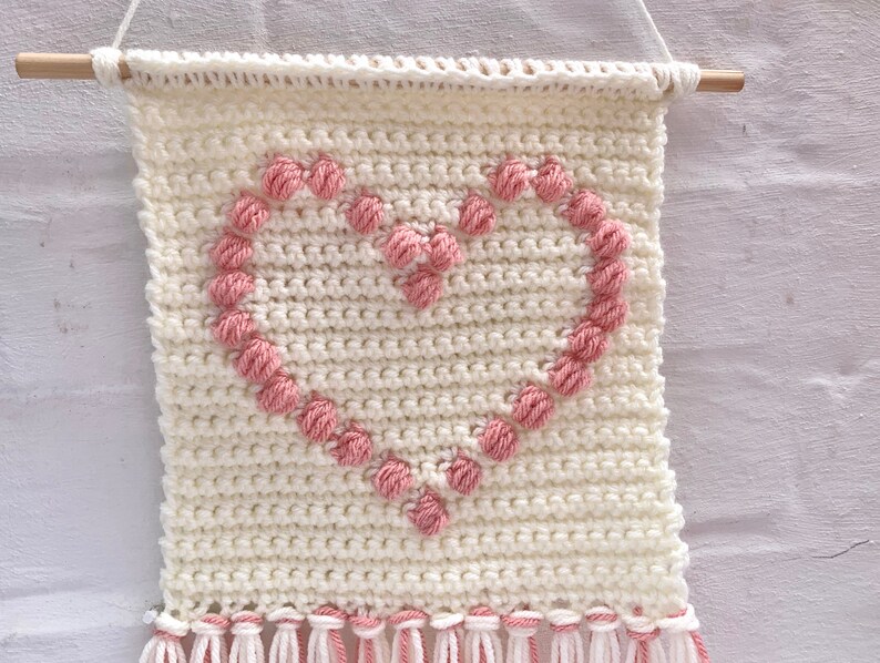 Op de afbeelding: Een witte gehaakte wandhanger met een roze hartontwerp. Het hart is gemaakt van kleine roze garenballen. De hanger heeft witte franjes aan de onderkant.