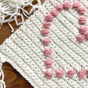 Op de afbeelding: Wit gecrochet vierkant met roze garen pompoenen die een hartvorm vormen.