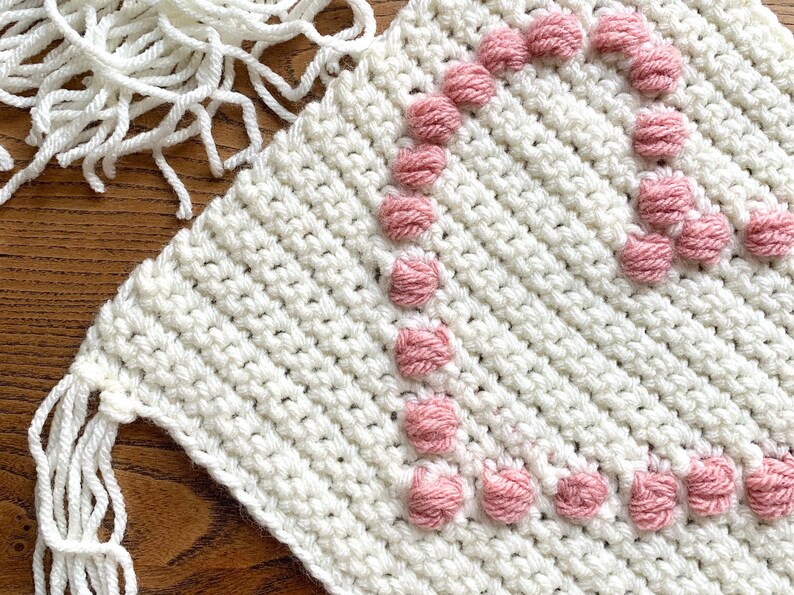 Op de afbeelding: Wit gecrochet vierkant met roze garen pompoenen die een hartvorm vormen.
