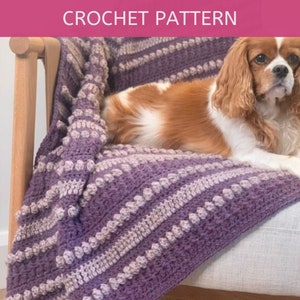 Può includere: Una coperta all'uncinetto viola e beige chiaro, drappeggiata su una sedia. La coperta presenta un motivo strutturato con bobi e strisce. Il testo "CROCHET PATTERN" è visualizzato in alto.