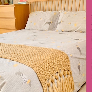 May include: A tan crocheted throw blanket with fringe on a bed with a white and grey patterned duvet cover. The bed has a wooden headboard with a light wood finish.