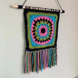 May include: A colorful crocheted wall hanging with a geometric pattern in shades of pink, green, blue, and black. The hanging has a fringe border.