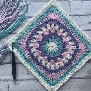 May include: A square crochet granny square with a white center and concentric rings of pink, purple, and teal yarn.