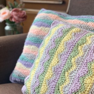 Puede incluir: Dos cojines de crochet con un patrón a rayas en tonos de rosa, amarillo, verde y morado.