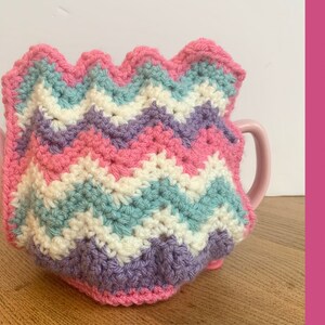 May include: A crocheted teapot cozy in pink, white, purple, and teal with a scalloped edge.