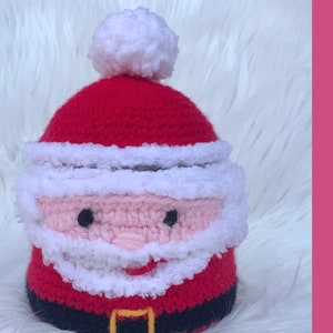 May include: Crocheted Santa Claus ornament with a red hat, white beard, and black belt. The ornament is made with pink and white yarn.