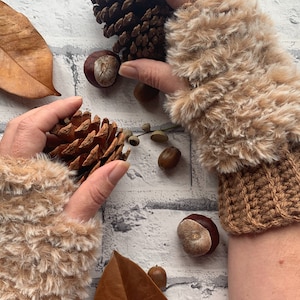 May include: A pair of brown and white fuzzy knit wrist warmers with a brown crocheted cuff. The wrist warmers are on a white brick background with pine cones and acorns.