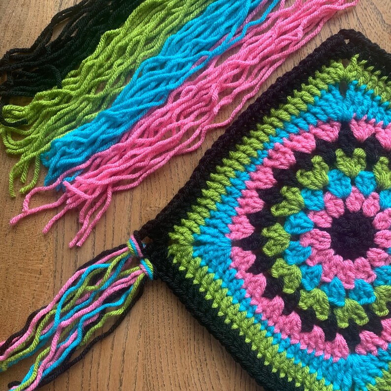 May include: A black, green, pink, and blue crocheted square with a fringe border. The fringe is made of multiple strands of yarn in pink, blue, green, and black.