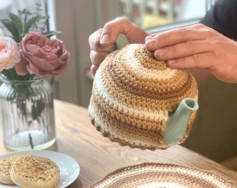 May include: A crocheted teapot cosy in brown and white stripes, being held by a person's hands. The cosy is on a teal teapot.