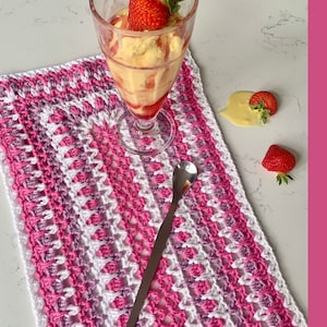 Puede incluir: Un mantel individual de crochet rosa y blanco con un borde festoneado. El mantel individual está colocado sobre una superficie blanca con un vaso de helado y fresas.