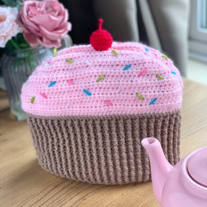 May include: A pink and brown crocheted tea cosy shaped like a cupcake with colourful sprinkles and a red cherry on top.