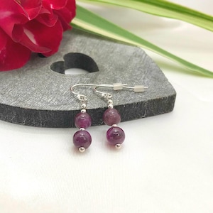 May include: A pair of silver dangle earrings with three purple gemstone beads. The earrings are displayed on a heart-shaped stone.