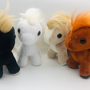 Könnte beinhalten: Vier Plüschpferde in Schwarz, Weiß, Beige und Orange. Jedes hat eine flauschige Mähne und einen Schweif in einer Kontrastfarbe. Die Pferde haben große, runde schwarze Augen und stehen auf einer weißen Oberfläche.