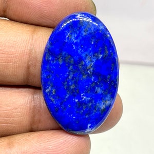 May include: Oval-shaped, polished lapis lazuli gemstone. The deep blue stone has flecks of gold and pyrite, creating a speckled appearance. The gemstone is held between fingers, showcasing its smooth, glossy surface.