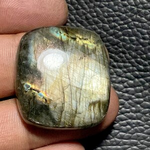 Puede incluir: Una piedra preciosa de labradorita de forma cuadrada con un brillo verde, azul y dorado. La piedra está pulida y tiene una superficie lisa.