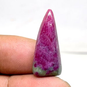 Puede incluir: Una gema pulida y alargada con un tono magenta intenso, que se estrecha hasta un punto. La piedra tiene una ligera base verde y una superficie lisa y reflectante. La gema se sostiene entre los dedos.