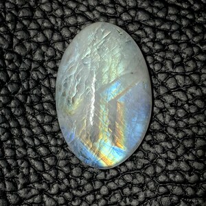 May include: Oval-shaped, iridescent moonstone gemstone. The stone displays a milky white base with flashes of blue, yellow, and orange. The polished surface reflects light, creating a shimmering effect. The gemstone is set against a dark, textured background.
