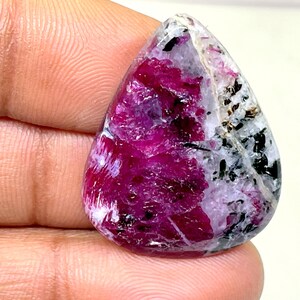 May include: A teardrop-shaped cabochon gemstone with a pink and white pattern. The gemstone has a dark grey vein running through it.