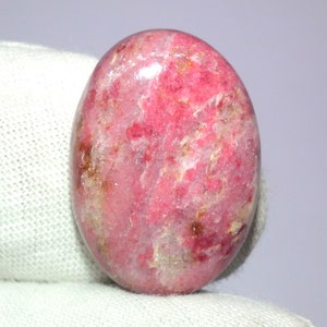 May include: A smooth, oval-shaped pink and white gemstone with a polished finish. The stone has a mottled appearance with pink and white swirls and flecks of brown.