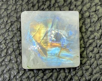 May include: A square, iridescent labradorite gemstone with a blue, green, and gold sheen. The stone is set in a clear, white, square cabochon.