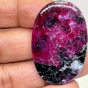 Puede incluir: Una gema ovalada de un vibrante color magenta, intercalada con inclusiones negras y blancas. La superficie pulida refleja la luz, mostrando los patrones y texturas naturales de la piedra. La gema es sostenida por una mano.