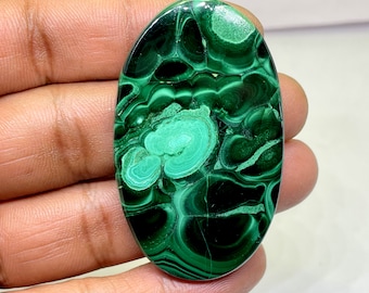 May include: Oval-shaped malachite gemstone with a polished surface. The stone features a swirling pattern of dark and light green hues, creating a marbled effect. The gemstone is held in a hand, showcasing its natural beauty and unique design.