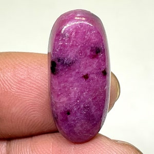 Könnte beinhalten: Ein rosafarbener ovaler Cabochon-Edelstein mit dunklen Einschlüssen. Der Edelstein ist poliert und hat eine glatte, glänzende Oberfläche.