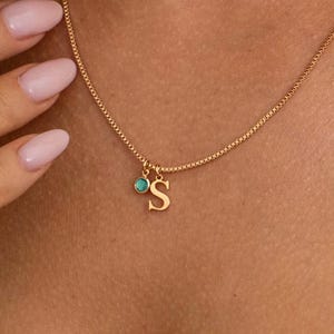 May include: A gold necklace with a fine chain. It has a small, gold letter "S" pendant and a small turquoise stone. The necklace is worn around the neck.