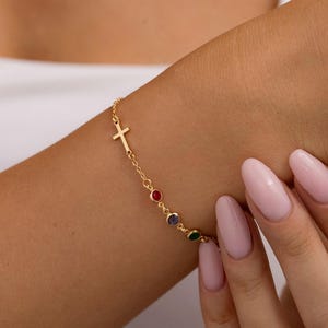 May include: Gold-toned bracelet featuring a small cross charm and colorful gemstones. The bracelet has a delicate chain design and is adorned with red, blue, and green stones. A stylish piece of jewelry.