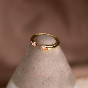 Può includere: Un anello aperto color oro con due estremità distinte. Un'estremità è decorata con una piccola pietra rotonda rosa chiaro, mentre l'altra estremità presenta una pietra più grande a forma di goccia, anch'essa di colore rosa chiaro.